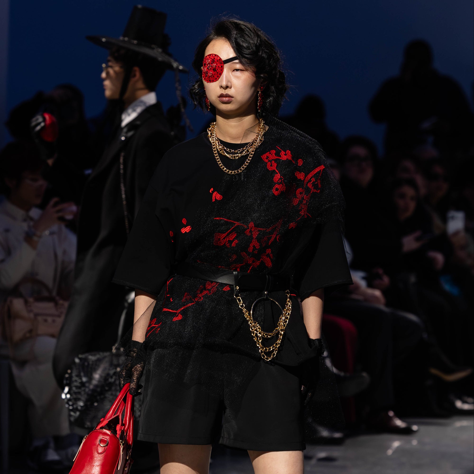 Model on a fashion runway wearing a black outfit with red embroiderd cherry blossom accents and holding a red bag.