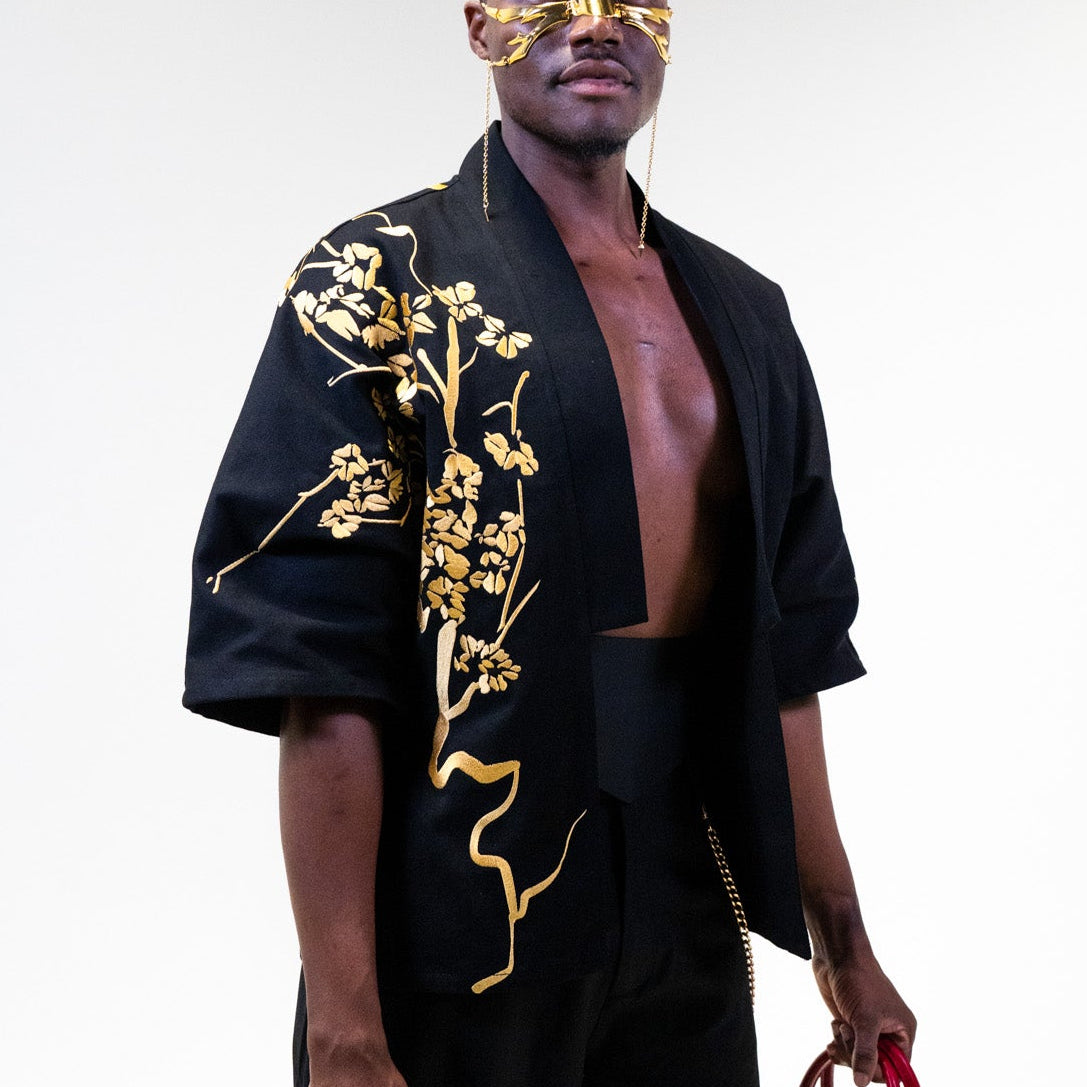 Man wearing a black limited edition Yelsh street Kimono outfit with gold embroidery, holding a red handbag against a white background